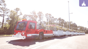 A red tram vehicle with multiple connected carriages is parked on a road, surrounded by trees under bright sunlight.