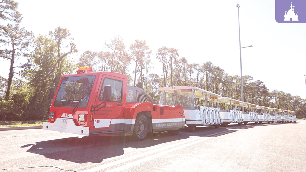 A red tram vehicle with multiple connected carriages is parked on a road, surrounded by trees under bright sunlight.