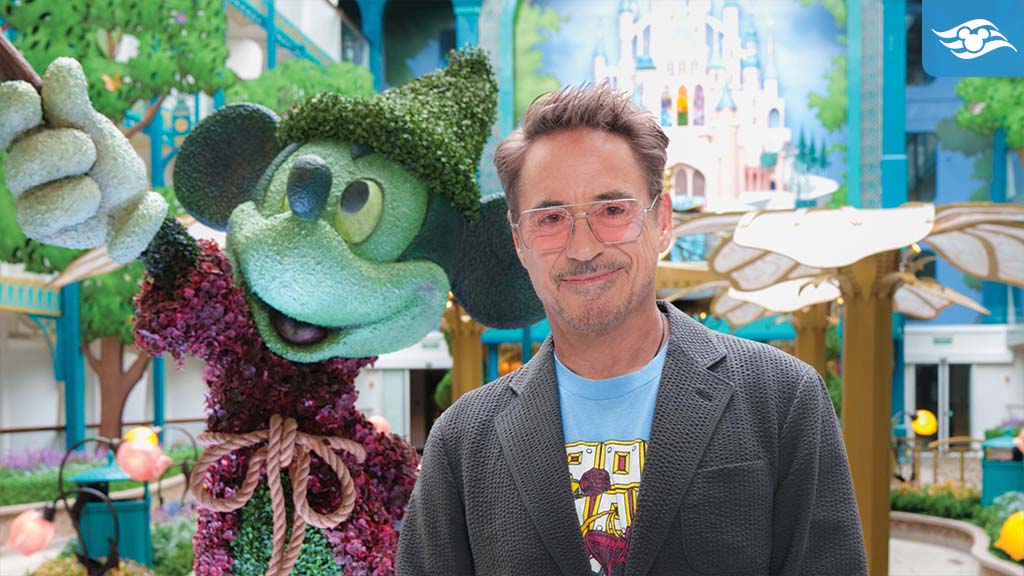 Robert Downey Jr. poses beside a Mickey Mouse topiary aboard Disney Adventure, with colorful Disney castle artwork in the background.