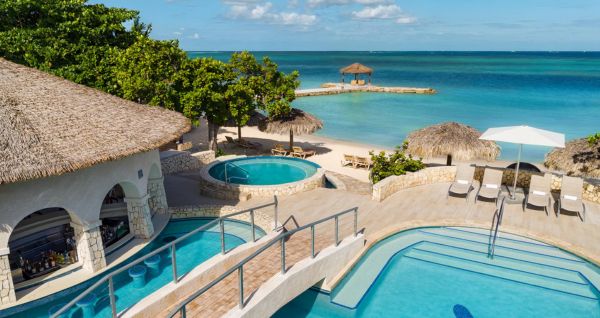 A tropical pool area at Sandals Montego Bay featuring a swim-up bar, a thatched-roof bridge, hot tub, lounge chairs, and a private beach overlooking the turquoise Caribbean Sea.