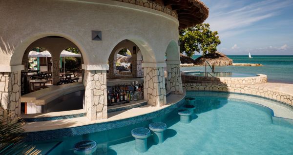 Swim-up bar at Sandals Montego Bay featuring stone archways, submerged barstools, and a scenic ocean view with a sailboat in the distance.