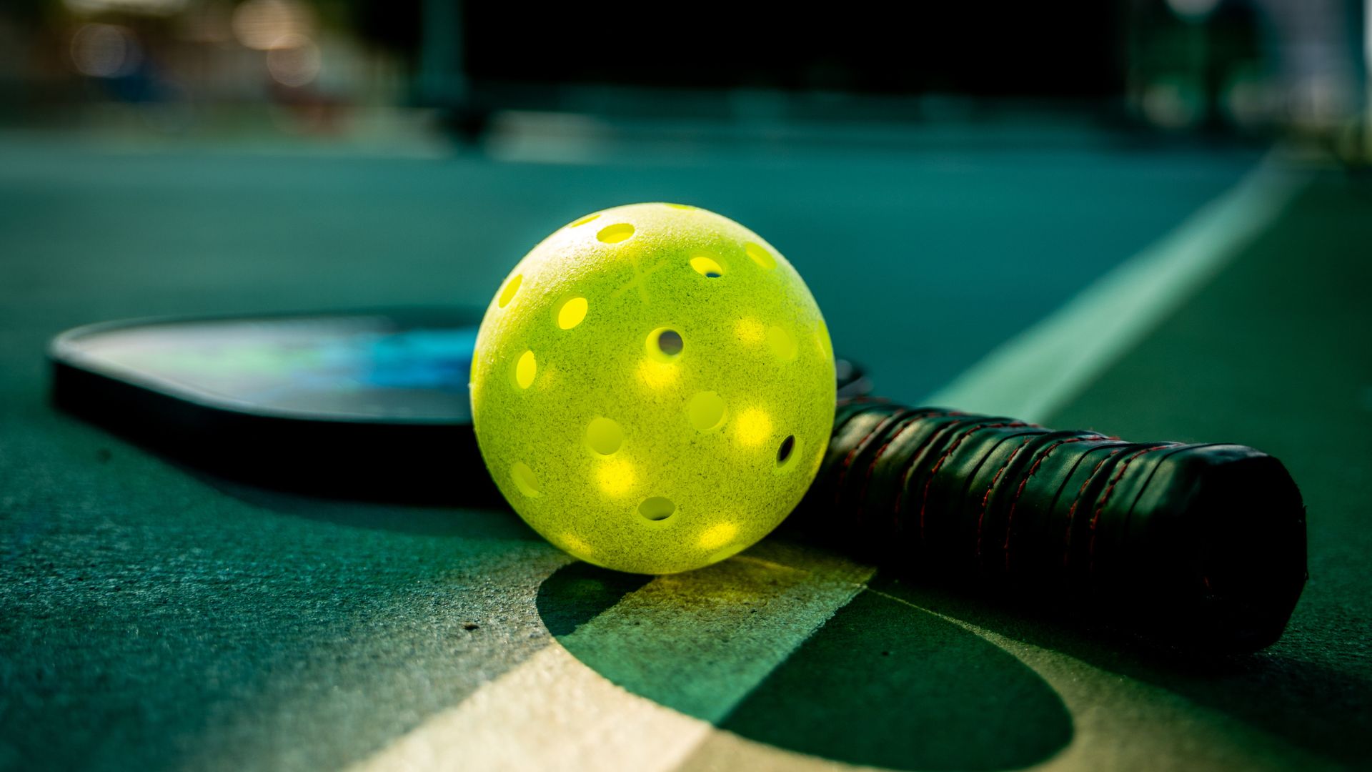 Close-up of a yellow pickleball and paddle lying on a court, with sunlight casting shadows.