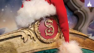 Santa hat resting on an ornate gold chair with a red "S" emblem, set against a festive Disney backdrop.