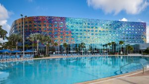 Large outdoor pool with lounge chairs, palm trees, and a colorful, oval-windowed hotel building in the background.