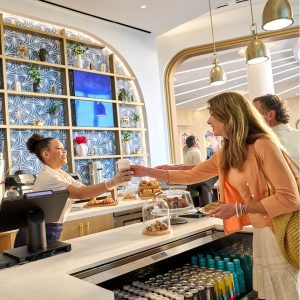 Smiling barista hands a coffee to a guest at Aurora Market inside Universal Helios Grand Hotel, with pastries and drinks on display.