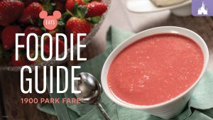 A bowl of bright pink soup sits on a green cloth, accompanied by a silver spoon and a bowl of fresh strawberries in the background.