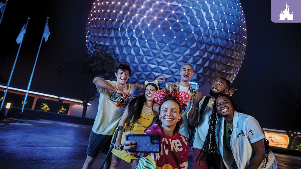 A group of six friends smiling and taking a selfie in front of the illuminated Epcot Spaceship Earth at night, wearing casual and Disney-themed clothing.