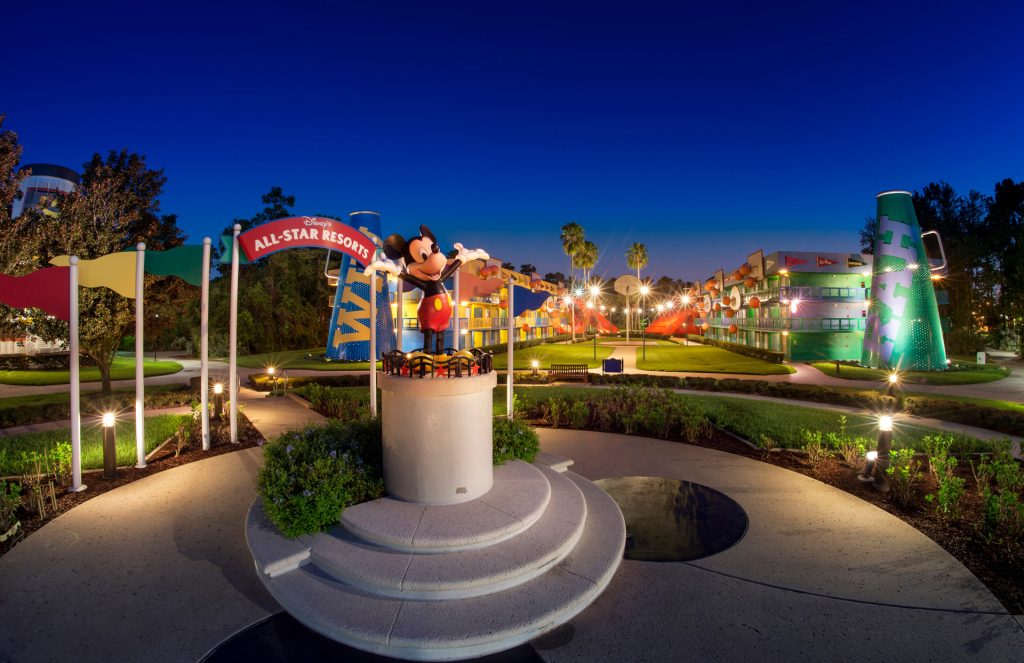 A colorful park scene at dusk features a statue of a character on a pedestal, with flags and festive lights illuminating the area.