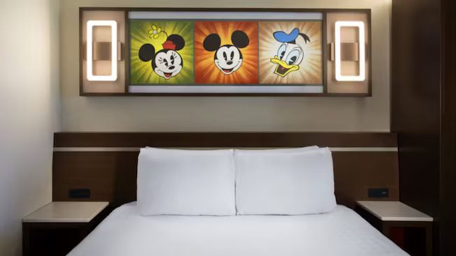 A cozy hotel room featuring a bed with white linens, adorned with a colorful wall art of Mickey Mouse and Donald Duck.