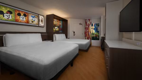 A modern hotel room features two beds, a large TV, colorful artwork on the walls, and a bright window with a view.