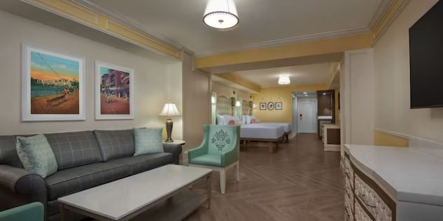 A cozy hotel room featuring a gray sofa, light wood furniture, colorful artwork, and a king-sized bed in the background.