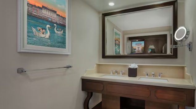 A clean bathroom with a wooden vanity, two sinks, a mirror, and a framed painting of swans on water. Light fixtures illuminate the space.