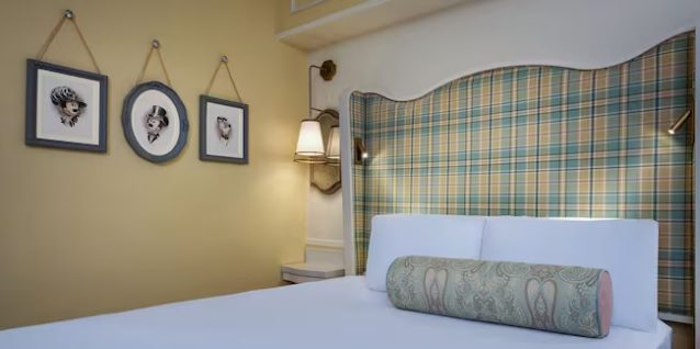 A cozy bedroom with a plaid headboard, white bedding, and three framed artworks on the wall. Soft lighting completes the inviting ambiance.