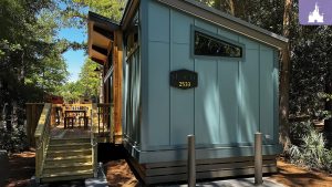 A small, blue cabin with a wooden deck, surrounded by trees. A table and chairs are visible on the deck. Number 2533 is displayed on the cabin.