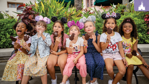 Seven young girls wearing Mickey ears enjoy Mickey-shaped ice cream bars while sitting on a bench at Walt Disney World.