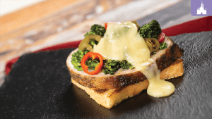 A toasted slice of bread topped with green vegetables, sliced red chili, and melted cheese, served on a black slate plate.
