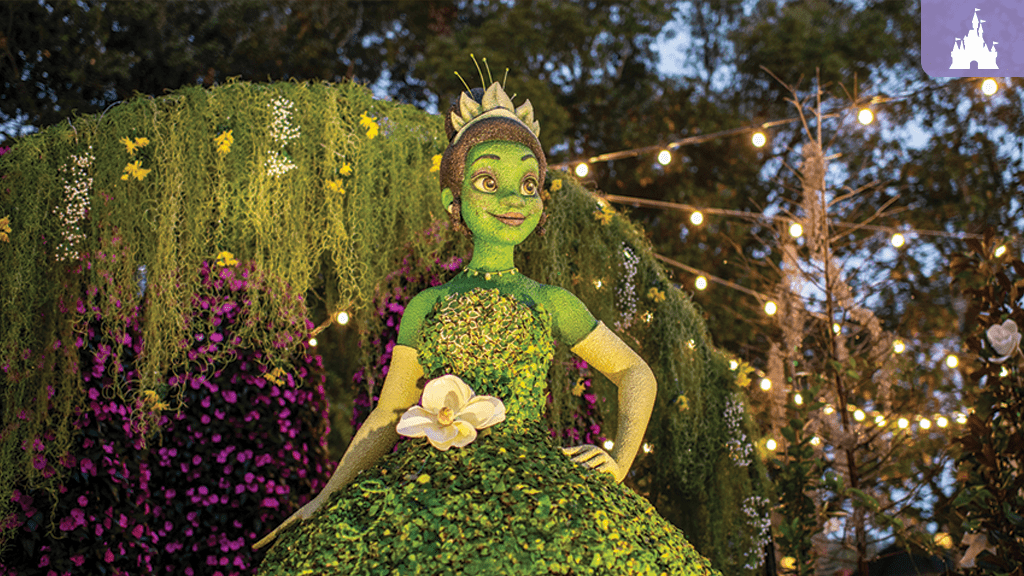 walt-disney-world-epcot-flower-garden-festival A character in a green dress adorned with plants and flowers stands amidst lush greenery and fairy lights, creating a magical atmosphere.