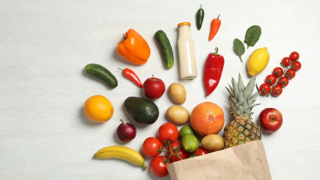 walt-disney-world-grocery-delivery Assorted fresh fruits and vegetables spilling from a paper grocery bag, including pineapple, tomatoes, bananas, peppers, avocados, and a bottle of milk on a white background.