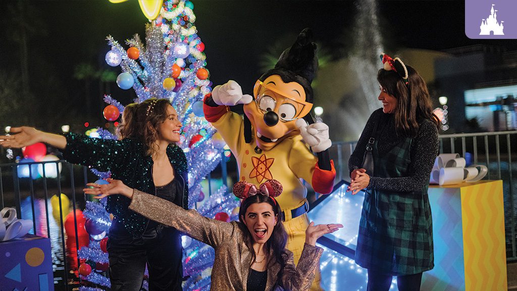 Three guests pose with Max Goof, dressed in his Powerline costume, in front of a festive Christmas tree decorated with colorful ornaments at Disney Jollywood Nights.
