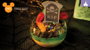 A dessert featuring crumbled cookies, gummy worms, colorful sprinkles, and a tombstone-shaped decoration labeled "RIP" in a clear cup.