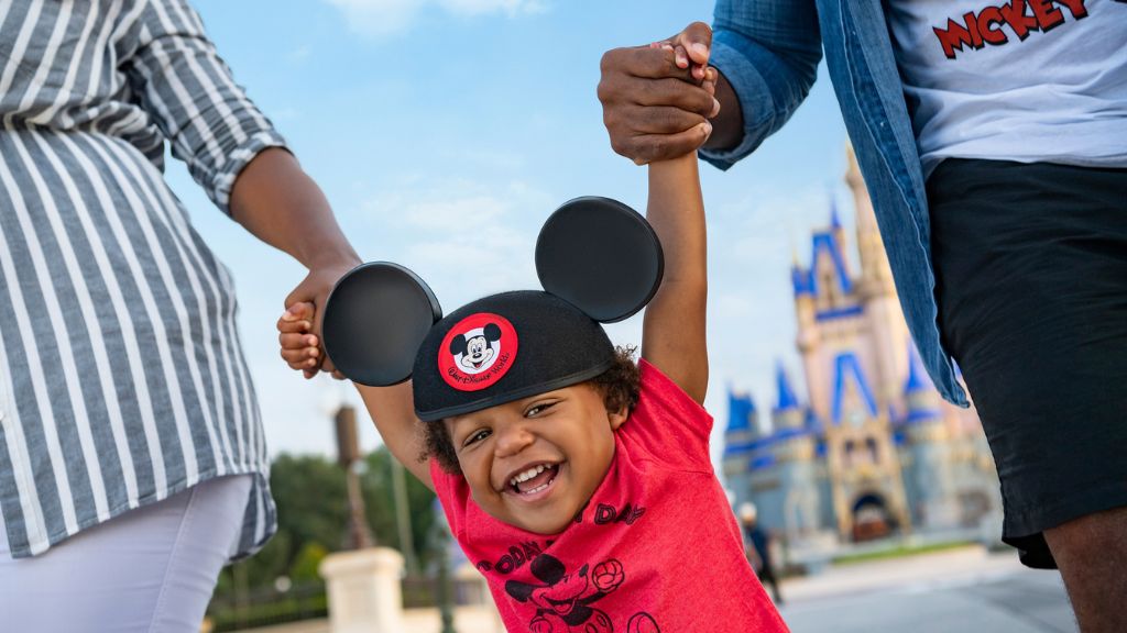 Smiling toddler wearing Mickey Mouse ears and a red Mickey shirt, holding hands with parents at Magic Kingdom in Walt Disney World with Cinderella Castle in the background.