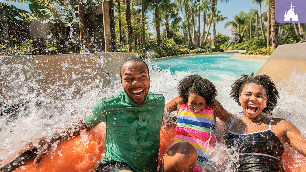 A man and two children smile joyfully as they splash down a waterslide, surrounded by lush greenery and a clear blue waterway.