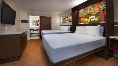 A cozy hotel room featuring two double beds, a flat-screen TV, and colorful wall art, with a modern design and wooden furnishings.