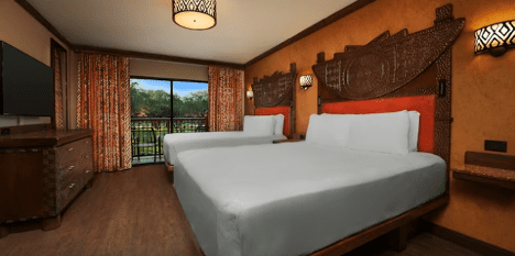 walt-disney-world-resort-animal-kingdom-lodge-one-bedroom-suite-bedroom A cozy hotel room featuring two queen beds, wooden decor, warm colors, and large windows with an outdoor view.