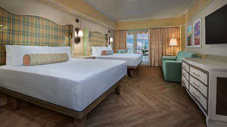 walt-disney-world-resort-boardwalk-inn-standard-room A bright hotel room featuring two beds, a large window with curtains, a dresser, and modern decor with warm wooden flooring.