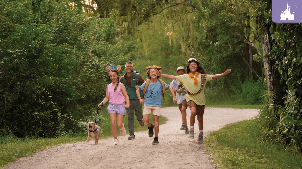 walt-disney-world-resort-cabins-fort-wilderness-family A group of five children and one adult walk playfully along a gravel path, surrounded by lush green trees, with a small dog leading the way.