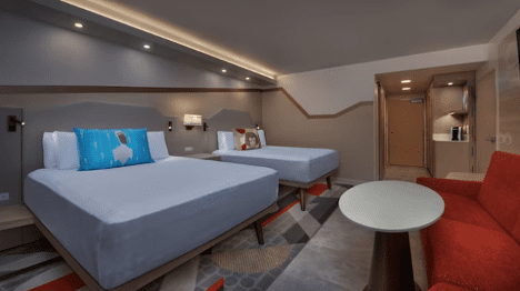 walt-disney-world-resort-contemporary-queen-room-beds A modern hotel room featuring two beds, a red couch, and a small table, with warm lighting and stylish decor.