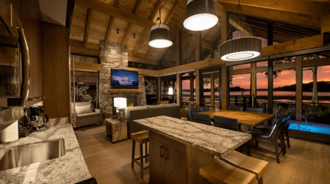 walt-disney-world-resort-copper-creek-cabin-interior Cozy modern kitchen and dining area with large windows showcasing a scenic sunset view. Wood beams and stone accents enhance the design.