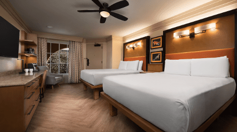 A cozy hotel room features two double beds, wooden furniture, a TV, and bright lighting, with curtains covering a window in the background.