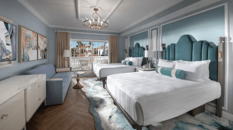 walt-disney-world-resort-grand-floridian-resort-studio A stylish hotel room with two beds, light blue walls, elegant furnishings, and a view of a sunny balcony area.