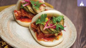Two bao buns filled with chicken, topped with avocado, pickled pink vegetables, and cilantro, served on a round plate.