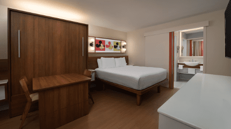 A cozy hotel room featuring a queen bed, wooden furniture, and a bright, modern design, with a bathroom visible in the background.
